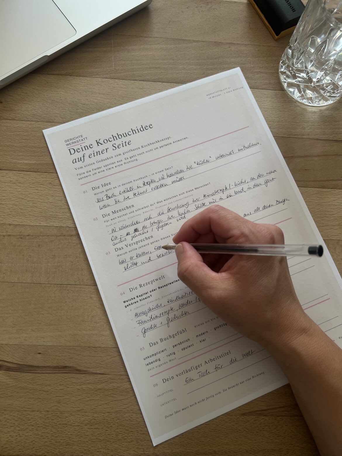 Spickzettel „Deine Kochbuchidee auf einer Seite“ mit Stift und Wasserglas